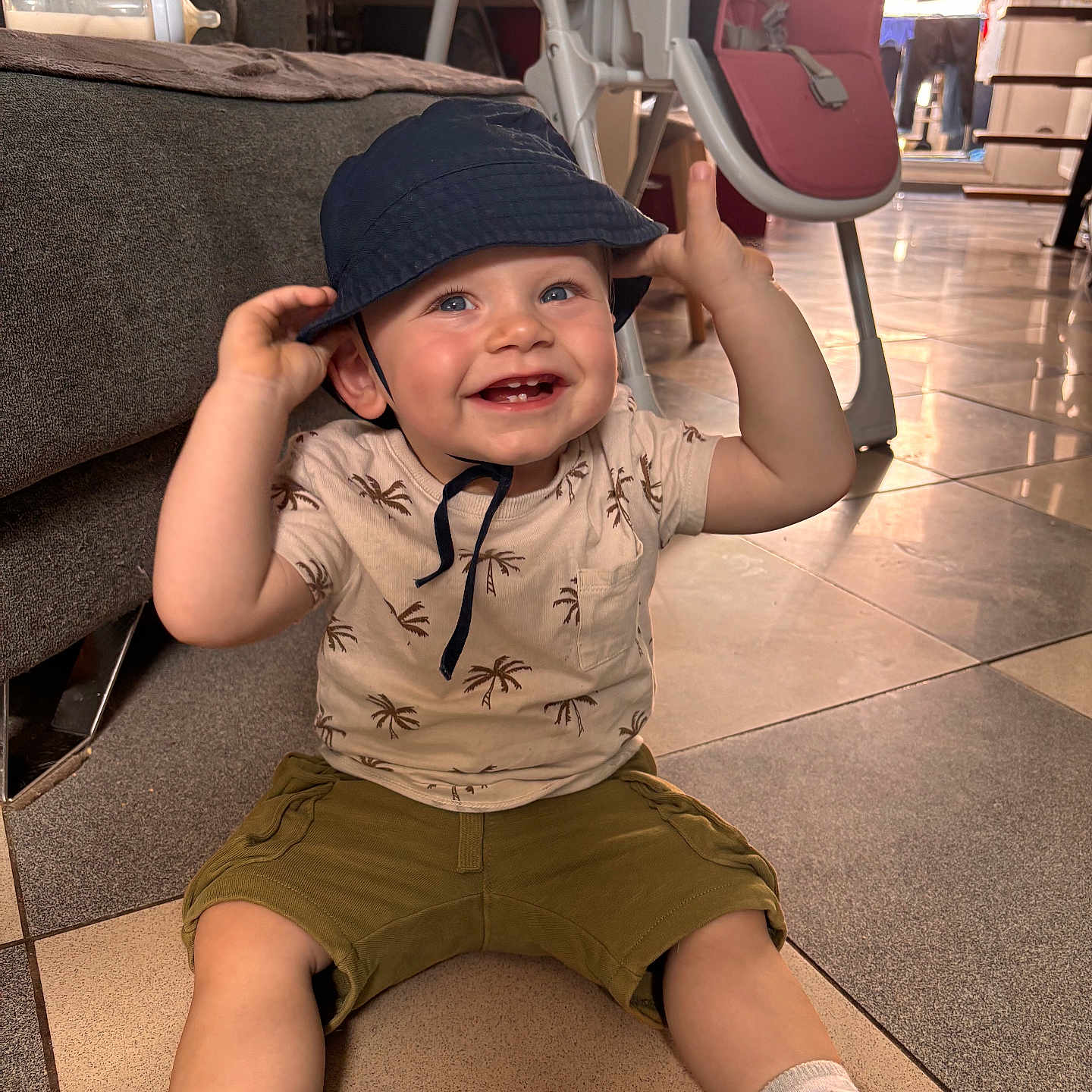 Andréas a rejoint le concours — aidez-le/la à gagner de superbes lots ! baby, baby_bottle, bucket_hat, casual_clothing, child, floor, footwear, happy, hat, high_chair, indoor, living_room, person, shirt, shorts, smiling, sneakers, sofa, tiled_floor, toddler