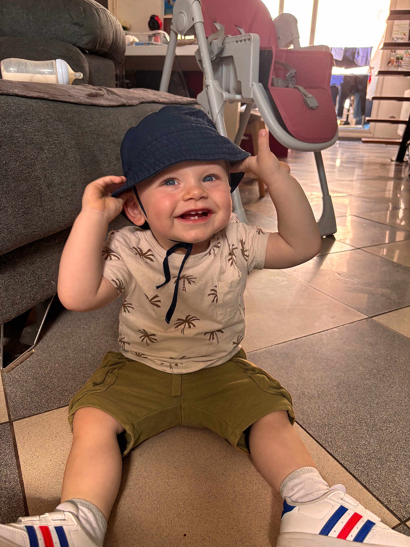 Andréas a rejoint le concours — aidez-le/la à gagner de superbes lots ! toddler, child, smiling, hat, bucket_hat, indoor, floor, tiled_floor, sneakers, shorts, shirt, baby_bottle, sofa, high_chair, living_room, happy, person, footwear, casual_clothing, baby