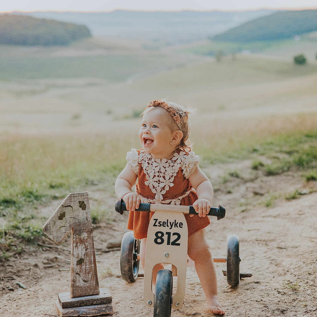Zselyke is registered to the contest to win money with this photo: barefoot, birthday, child, cute, dirt_path, dress, grass, happy, headband, hill, nature, number_one, outdoor, play, portrait, rustic, smiling, sunlight, toddler, wooden_tricycle
