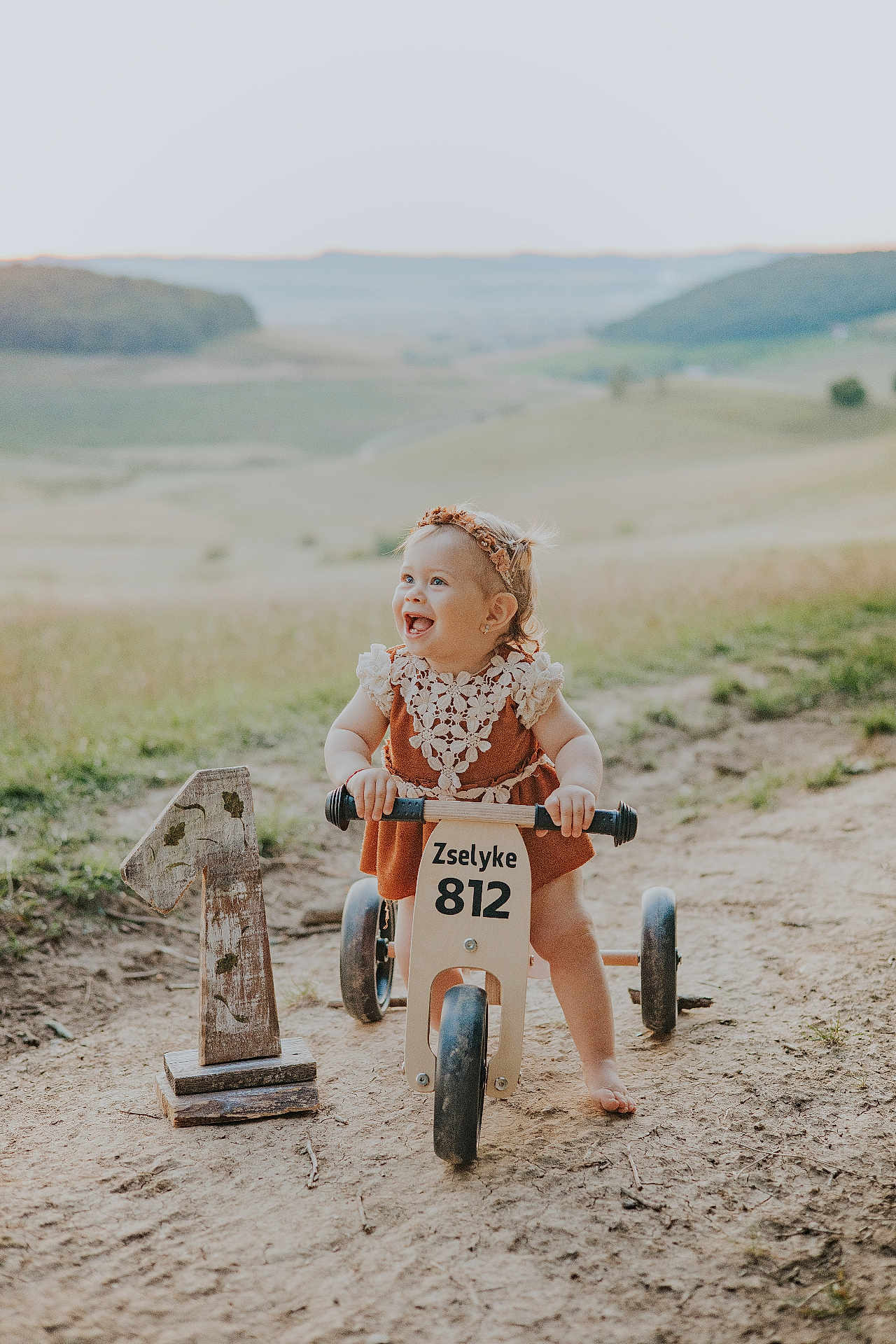 Zselyke is registered to the contest to win money with this photo: toddler, child, smiling, outdoor, dirt_path, wooden_tricycle, headband, dress, number_one, birthday, nature, grass, hill, happy, portrait, sunlight, barefoot, play, cute, rustic