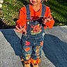 child, toddler, overalls, orange_shirt, hair_braids, sidewalk, grass, garden_hose, sunlight, shadow, playful, outdoor, smiling, cute, denim, fall_leaves, boots, person, portrait, daytime