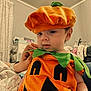 baby, child, costume, pumpkin, orange, hat, green_leaves, face, indoor, person, cute, portrait, hand, clothing, smile, baby_clothes, infant, soft_texture, cozy, room