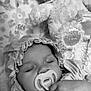 baby, sleeping, pacifier, bonnet, dress, teddy_bear, soft_toy, blanket, floral_pattern, close_up, portrait, child, infant, cute, peaceful, black_and_white, hand, cozy, resting, indoors