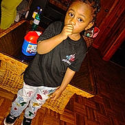 Khani is registered to the contest to win money with this photo: black_tshirt, casual_clothing, child, colorful_pants, curious_expression, door, flooring, hairstyle, home_interior, household_items, indoor, parquet_floor, person, plastic_bottle, sneakers, standing, thumb_in_mouth, wicker_table, wooden_walls, young_child