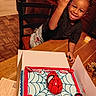 Khani is registered to the contest to win money with this photo: birthday_cake, black_tshirt, box, braided_hair, cake, celebration, chair, child, dessert, food, hand_gesture, happy, home, indoor, party, person, smiling, spiderman, table, wooden_floor