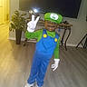 blue_overalls, casual_shoes, child, costume, decoration, face_paint, furniture, green_hat, home, indoor, living_room, luigi, peace_sign, plant, pose, smile, television, white_gloves, wooden_floor, young_boy