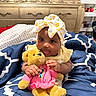 baby, child, teddy_bear, plush_toy, headwrap, blanket, bedroom, furniture, dresser, smiling, cute, indoors, clothing, infant, person, comfort, soft_toy, patterned, yellow, pink