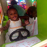 Alayah is registered to the contest to win money with this photo: toddler, child, arcade, kiddie_ride, steering_wheel, smiling, reflection, dress, white_dress, indoor, play, fun, colorful, toy_car, happy, young_child, amusement, entertainment, face, person