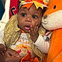 Alayah joined the competition — help win amazing prizes! baby, child, stuffed_animal, orange, headband, clothing, golden_vest, cute, hand, adult_hand, surprised_expression, indoors, portrait, soft_toy, toddler, closeup, face, expression, warm_colors, holding