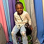 Khalani is registered to the contest to win money with this photo: child, girl, smiling, sitting, stool, jeans, shirt, hairstyle, braids, colorful_background, fabric, footwear, sneakers, purse, accessory, happy, portrait, indoor, playful, casual