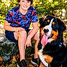 boy, child, dog, bernese_mountain_dog, pet, portrait, outdoor, nature, forest, trees, sunlight, smiling, seated, shorts, t_shirt, sneakers, long_tongue, floppy_ears, fallen_leaves, companion
