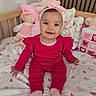 baby, child, pink_clothing, headband, smile, bed, bedspread, heart_pattern, plush_toys, doll, cushion, crib, indoor, person, infant, happy, cute, portrait, sitting, soft_toys
