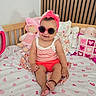Adalyn is registered to the contest to win money with this photo: baby, child, sunglasses, headband, bed, heart_pattern, stuffed_animals, pillow, pink, smile, cute, indoors, clothing, infant, person, playful, happy, toy, wooden_bedframe, colorful