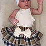 baby, infant, child, plaid_skirt, bow, headband, white_top, barefoot, lying_down, beige_background, cute_clothing, small_hands, curious_expression, indoors, soft_lighting, portrait, skin, face, person, toddler
