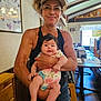 woman, baby, child, indoor, restaurant, glasses, visor, holding, smile, floral_onesie, wooden_furniture, poster, table, chair, natural_light, person, portrait, happy, casual_clothing, family