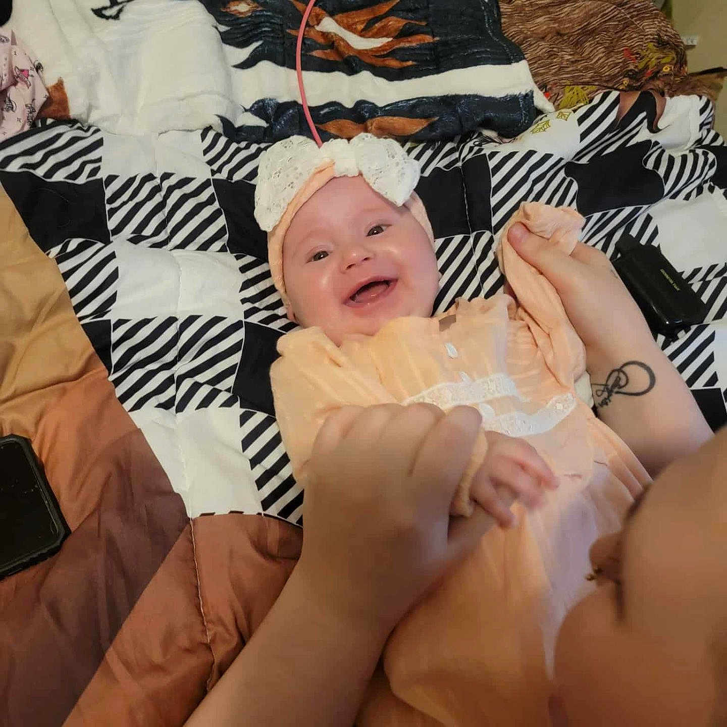 Skylynn joined the competition — help win amazing prizes! baby, bed, blanket, bow, child, closeup, face, hands, happy, headband, holding, indoor, infant, joy, orange_clothing, person, playful, quilt, smiling, tattoo
