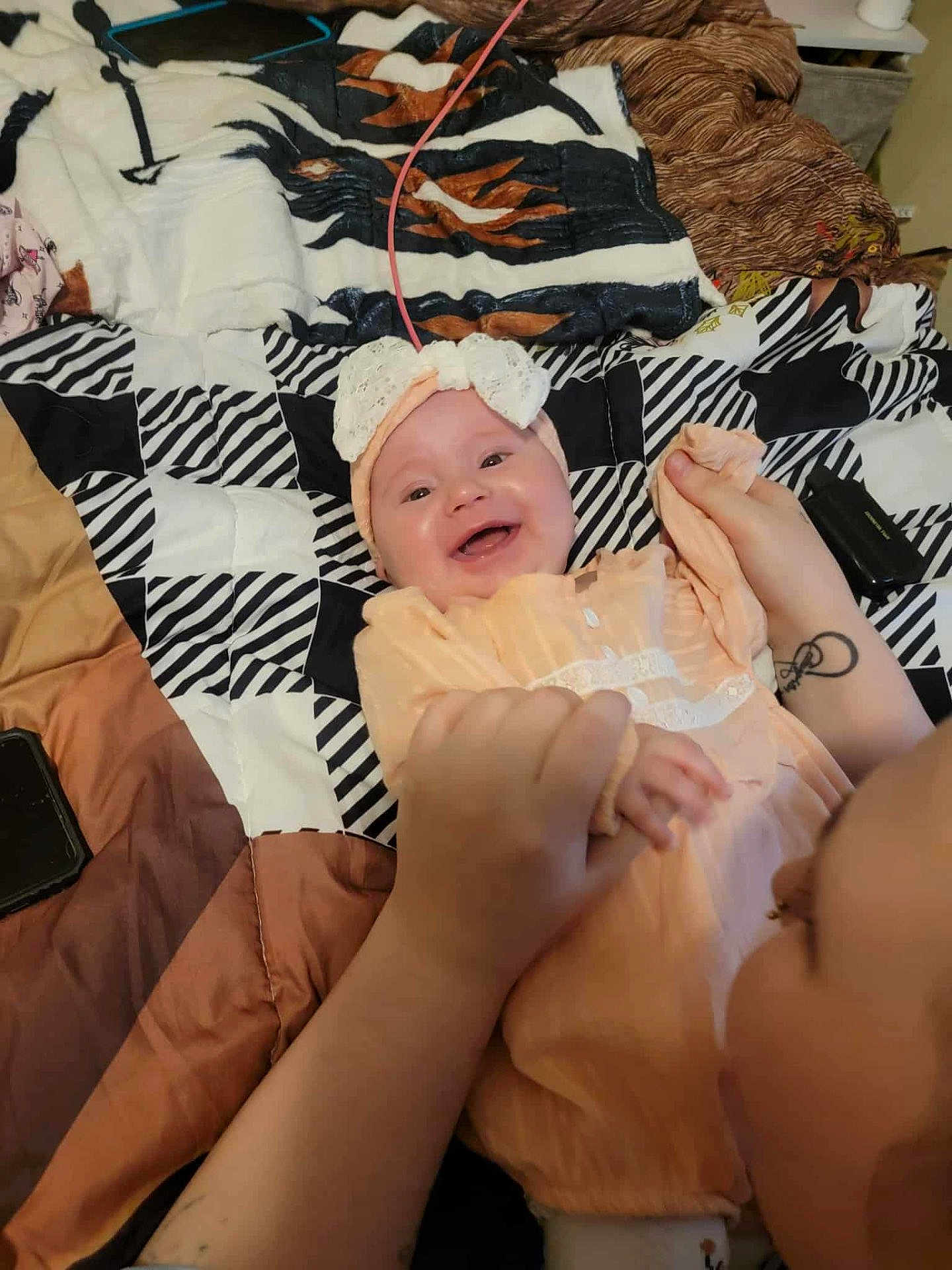 Skylynn joined the competition — help win amazing prizes! baby, child, smiling, happy, person, hands, headband, bow, quilt, blanket, bed, orange_clothing, tattoo, indoor, playful, closeup, face, infant, holding, joy