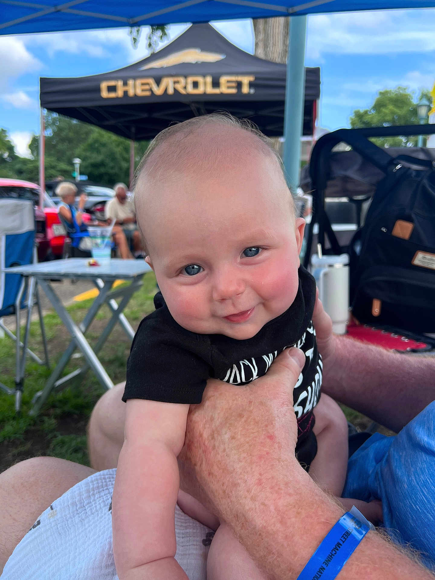 Grayson is registered to the contest to win money with this photo: baby, child, smiling, face, outdoor, people, hand, canopy, chevrolet, event, grass, table, chair, backpack, blue_sky, adult, person, clothing, holding, park