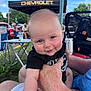 Grayson is registered to the contest to win money with this photo: baby, child, smiling, face, outdoor, people, hand, canopy, chevrolet, event, grass, table, chair, backpack, blue_sky, adult, person, clothing, holding, park