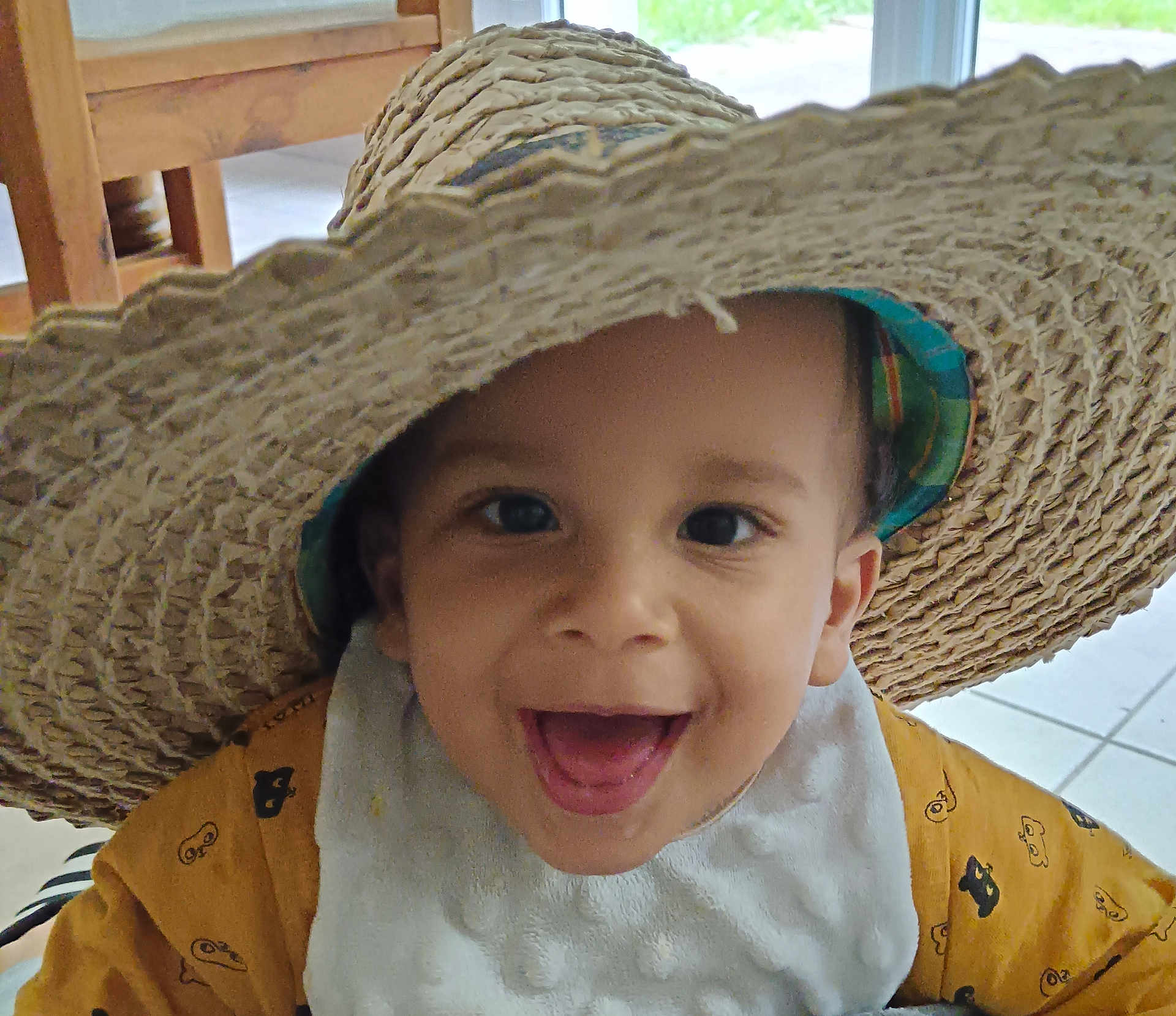 Tilyann'H a rejoint le concours — aidez-le/la à gagner de superbes lots ! toddler, child, smiling, hat, straw_hat, indoor, bib, happy, face, person, yellow_clothing, wooden_chair, floor, tile_floor, closeup, portrait, cute, baby, headwear, expression