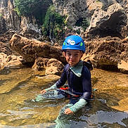 Lyham participe au concours pour gagner de l'argent avec cette photo : child, water, helmet, wetsuit, rocks, nature, outdoor, adventure, young, exploration, shallow_water, greenery, person, sitting, calm, sunlight, recreation, fun, river, water_sport