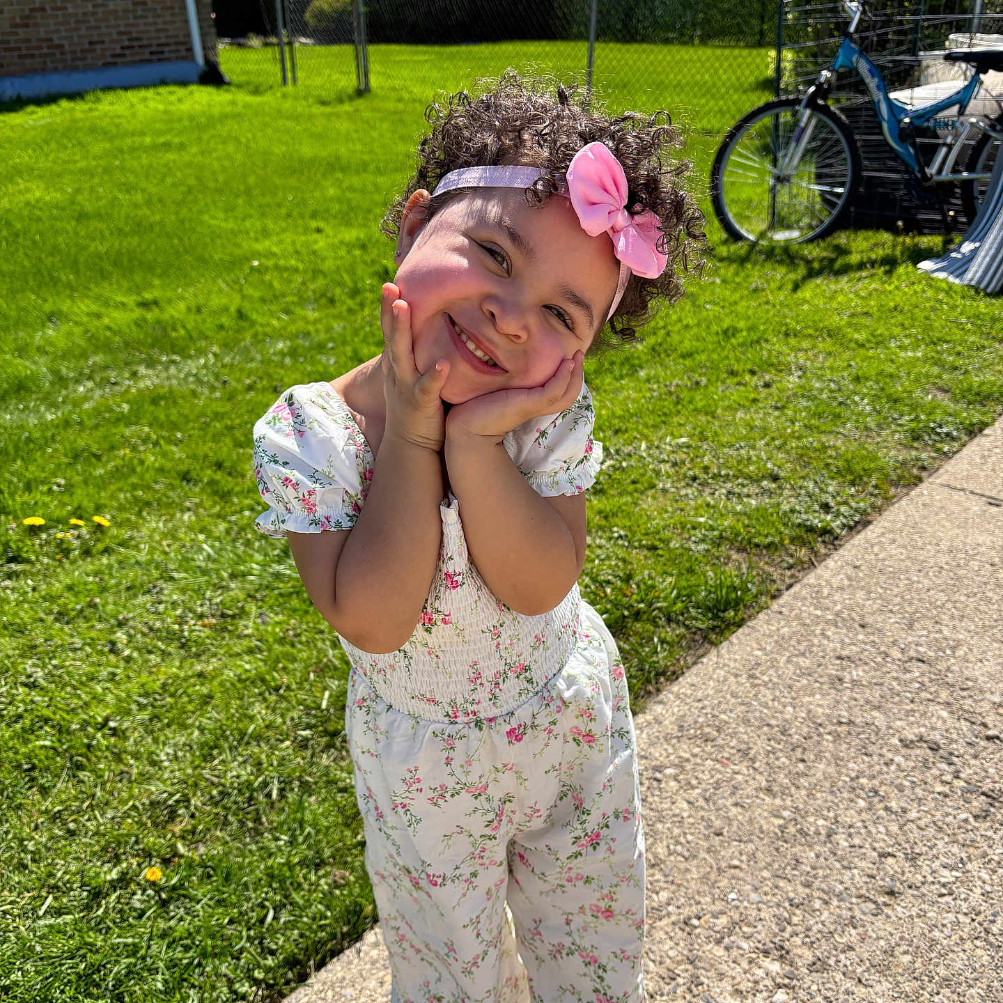 Isabella is registered to the contest to win money with this photo: bicycle, child, curly_hair, cute, daytime, flower_pattern, girl, grass, happy, headband, outdoor, pink_bow, playful, portrait, posing, sidewalk, smiling, sunny, white_clothing, young