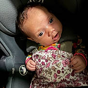 Hazel is registered to the contest to win money with this photo: baby, infant, child, car_seat, car_interior, harness, blanket, floral_pattern, wide_eyes, portrait, sitting, travel, vehicle, head, face, hands, clothing, indoor, close_up, safe