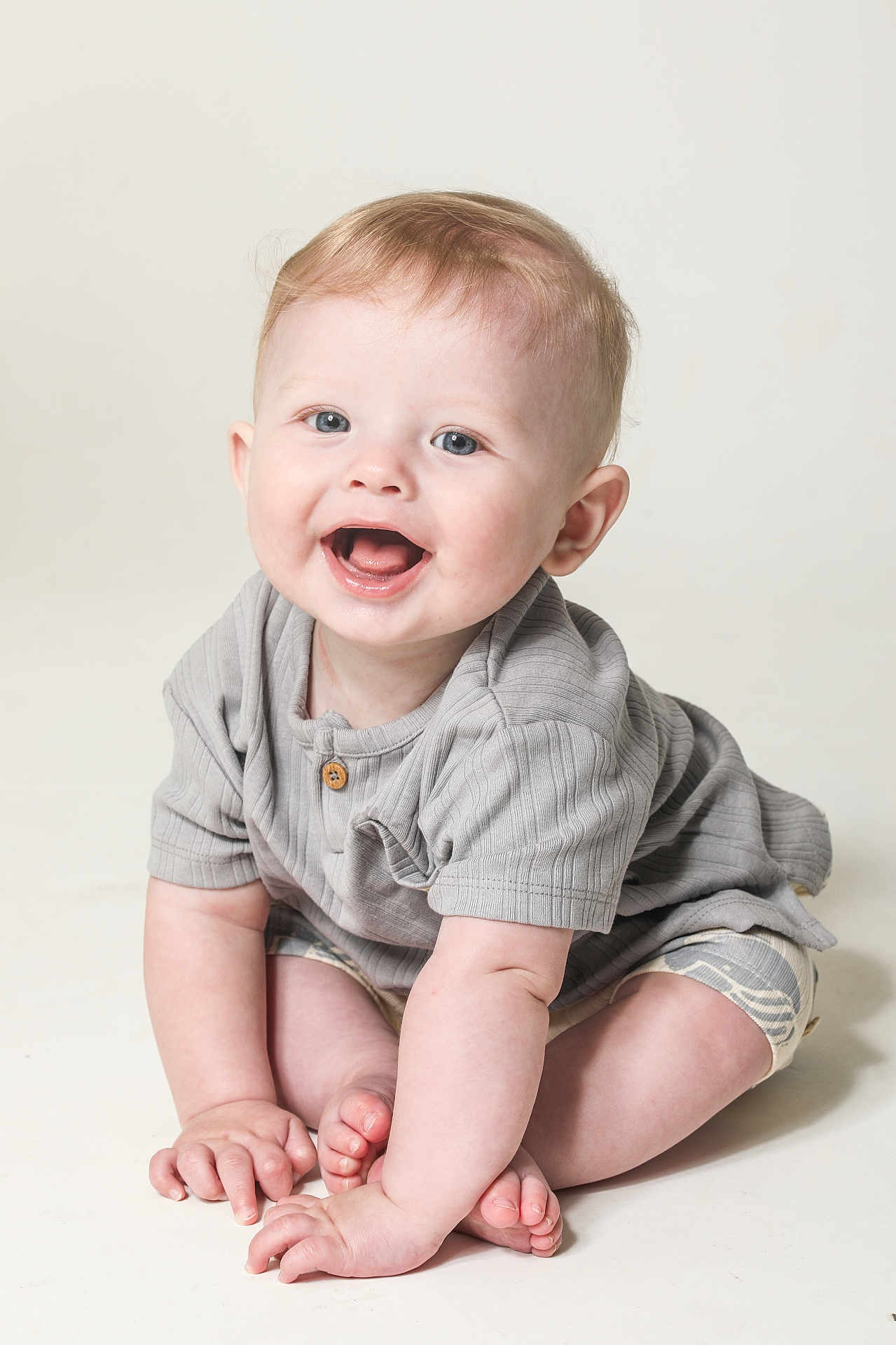 Rex is registered to the contest to win money with this photo: baby, child, smiling, sitting, happy, blue_eyes, shorts, shirt, cute, infant, hands, feet, skin, light, portrait, indoors, neutral_background, young, person, baby_clothes