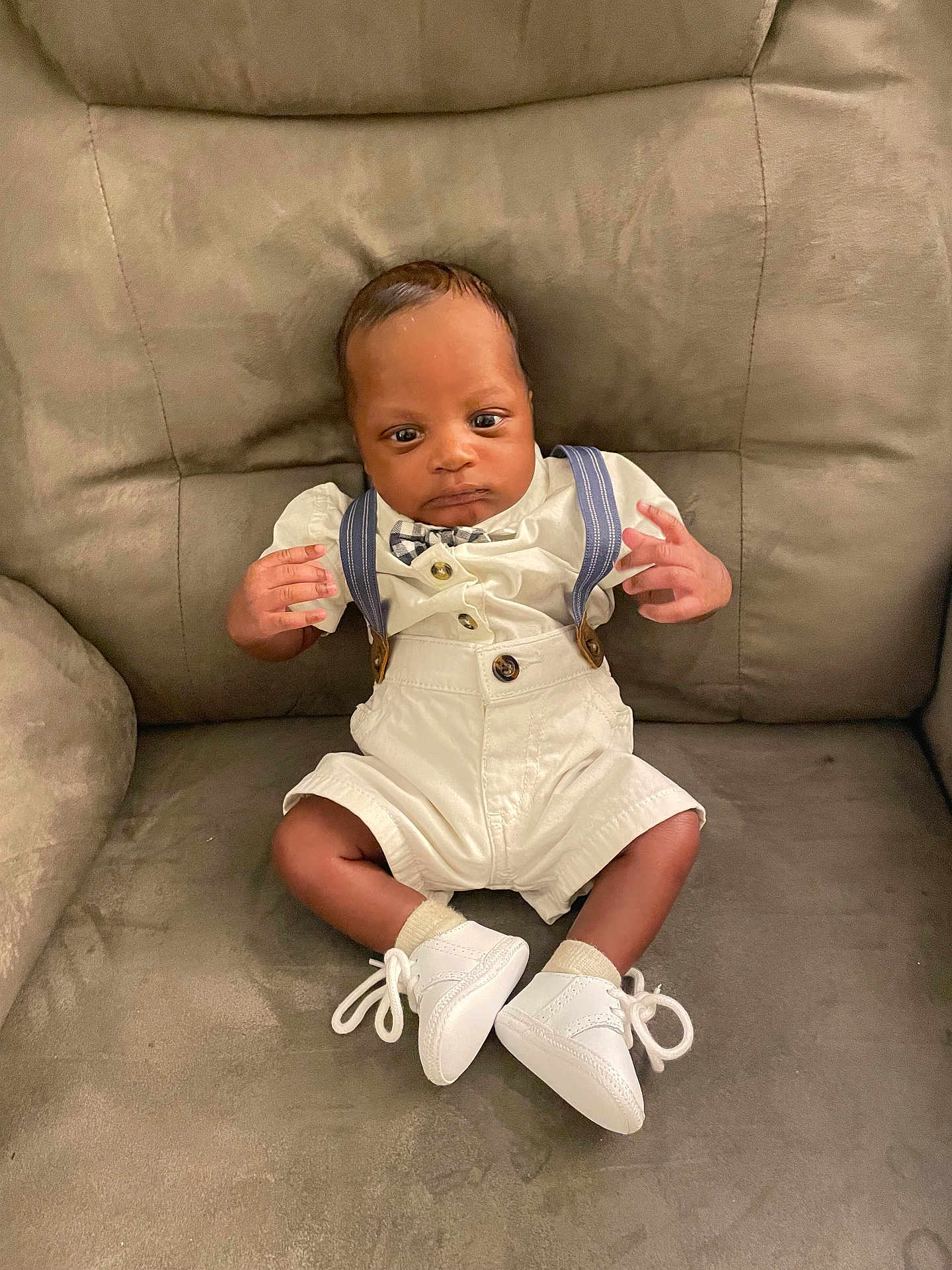 Roman is registered to the contest to win money with this photo: baby, infant, sitting, white_clothing, suspenders, bow_tie, white_shoes, couch, furniture, indoor, cute, child, portrait, small_hands, curious, young, person, fashion, comfortable, home
