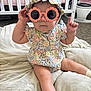 baby, child, sunglasses, floral_clothing, bonnet, romper, cushion, nursery, crib, carpet, sock, seated, indoor, cute, fashion, infant, person, playful, accessory, portrait