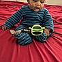 baby, bed, blue_onesie, child, curly_hair, curtains, expression, footwear, furniture, hand, indoor, infant, person, red_bedspread, remote_control, room, sitting, striped_clothing, toy, window