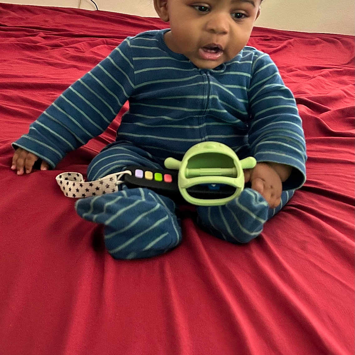 Kimani is registered to the contest to win money with this photo: baby, bed, blue_onesie, child, curly_hair, curtains, expression, footwear, furniture, hand, indoor, infant, person, red_bedspread, remote_control, room, sitting, striped_clothing, toy, window