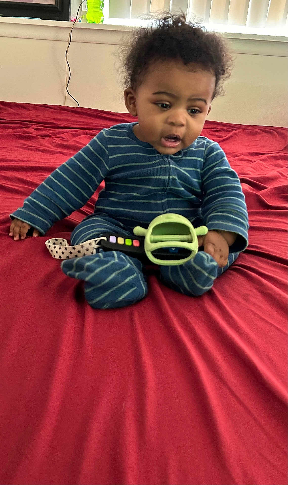 Kimani is registered to the contest to win money with this photo: baby, bed, blue_onesie, child, curly_hair, curtains, expression, footwear, furniture, hand, indoor, infant, person, red_bedspread, remote_control, room, sitting, striped_clothing, toy, window