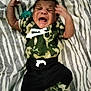 baby, infant, crying, camouflage, nike, clothing, blanket, pattern, zebra_stripes, laying_down, person, young_child, expression, indoor, apparel, footwear, sock, cute, hands, face