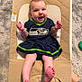Kennedy is registered to the contest to win money with this photo: baby, infant, smiling, seattle_seahawks, dress, baby_seat, cushion, carpet, toy, happy, legs, feet, indoor, person, child, cute, playful, clothing, skin, expression