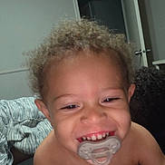 Ezekiel joined the competition — help win amazing prizes! toddler, child, smiling, curly_hair, pacifier, indoor, blanket, open_door, happy, person, face, mouth, teeth, skin, head, hair, baby, expression, bedroom, furniture