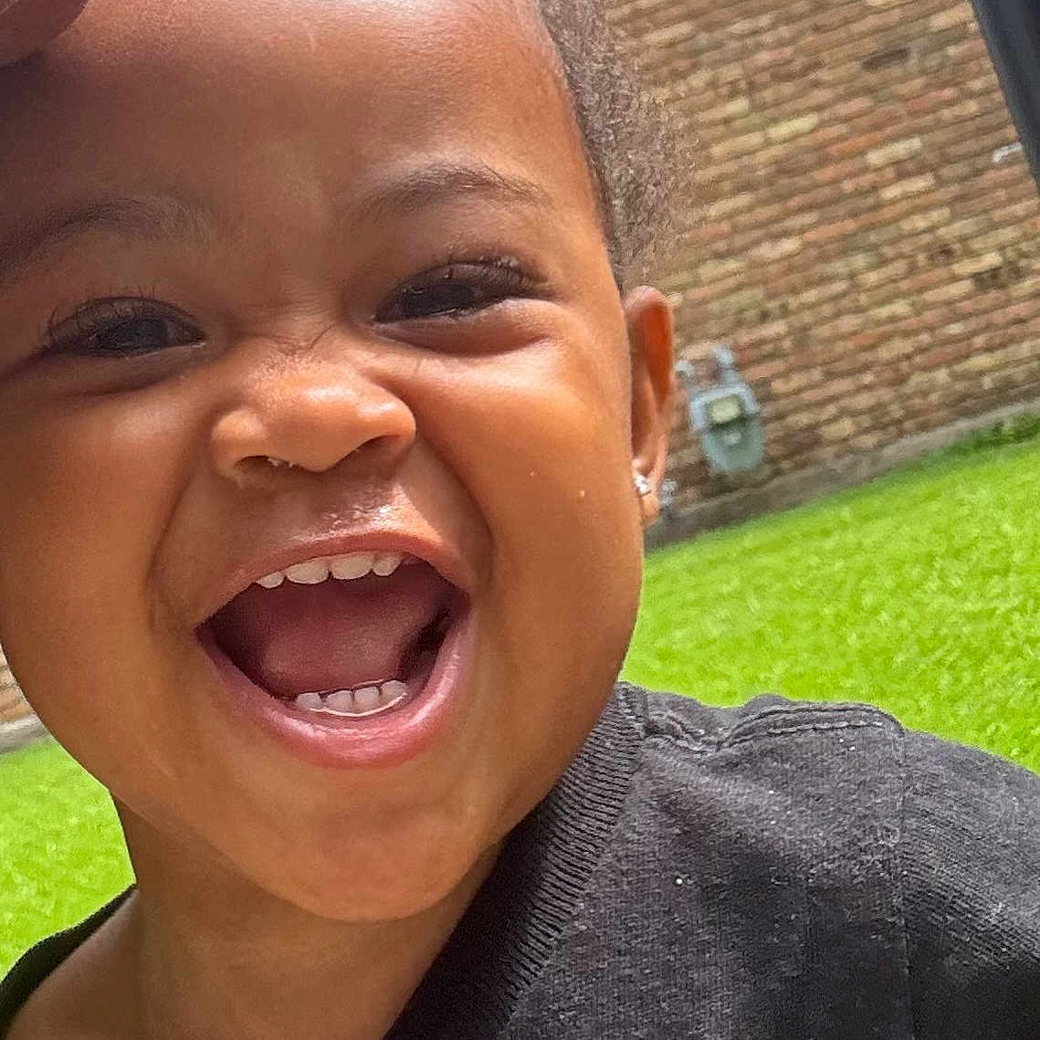 Janae is registered to the contest to win money with this photo: black_clothing, brick_wall, child, closeup, earring, expression, eye, face, grass, hair, happy, head, joy, mouth, outdoor, person, skin, smiling, teeth, toddler