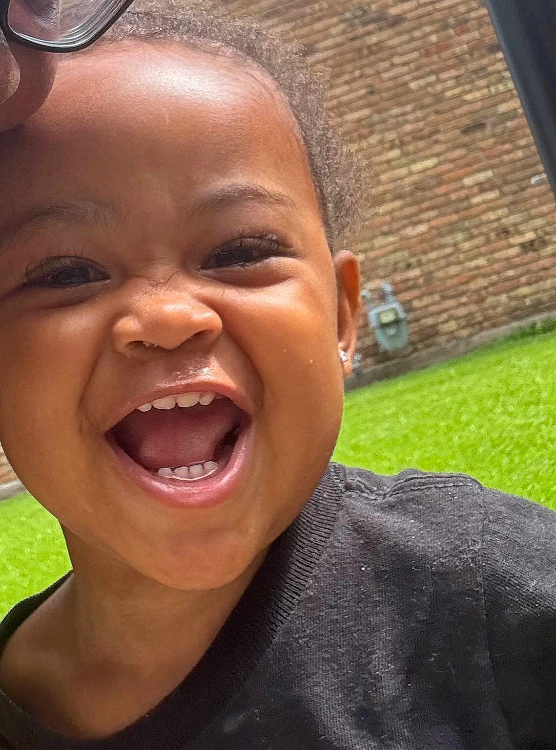 Janae is registered to the contest to win money with this photo: child, toddler, smiling, happy, face, teeth, black_clothing, outdoor, grass, brick_wall, earring, closeup, joy, expression, person, skin, head, mouth, eye, hair