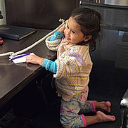 Celestia is registered to the contest to win money with this photo: toddler, child, telephone, office_chair, desk, sweater, pajamas, smile, indoor, curly_hair, coiled_cord, notebook, pen, black_chair, wooden_desk, person, cute, playful, communication, home_office