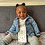 baby, child, smiling, denim_jacket, pants, socks, face, hair, couch, sign, text, indoor, happy, cute, infant, sitting, carpet, wall, person, portrait