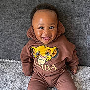 Noah is registered to the contest to win money with this photo: baby, child, smiling, hoodie, brown_clothing, sitting, carpet, couch, indoor, cute, portrait, happy, face, person, young_child, casual_wear, cartoon_graphic, lion_king, comfortable, cozy