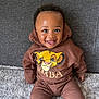 baby, child, smiling, hoodie, brown_clothing, sitting, carpet, couch, indoor, cute, portrait, happy, face, person, young_child, casual_wear, cartoon_graphic, lion_king, comfortable, cozy