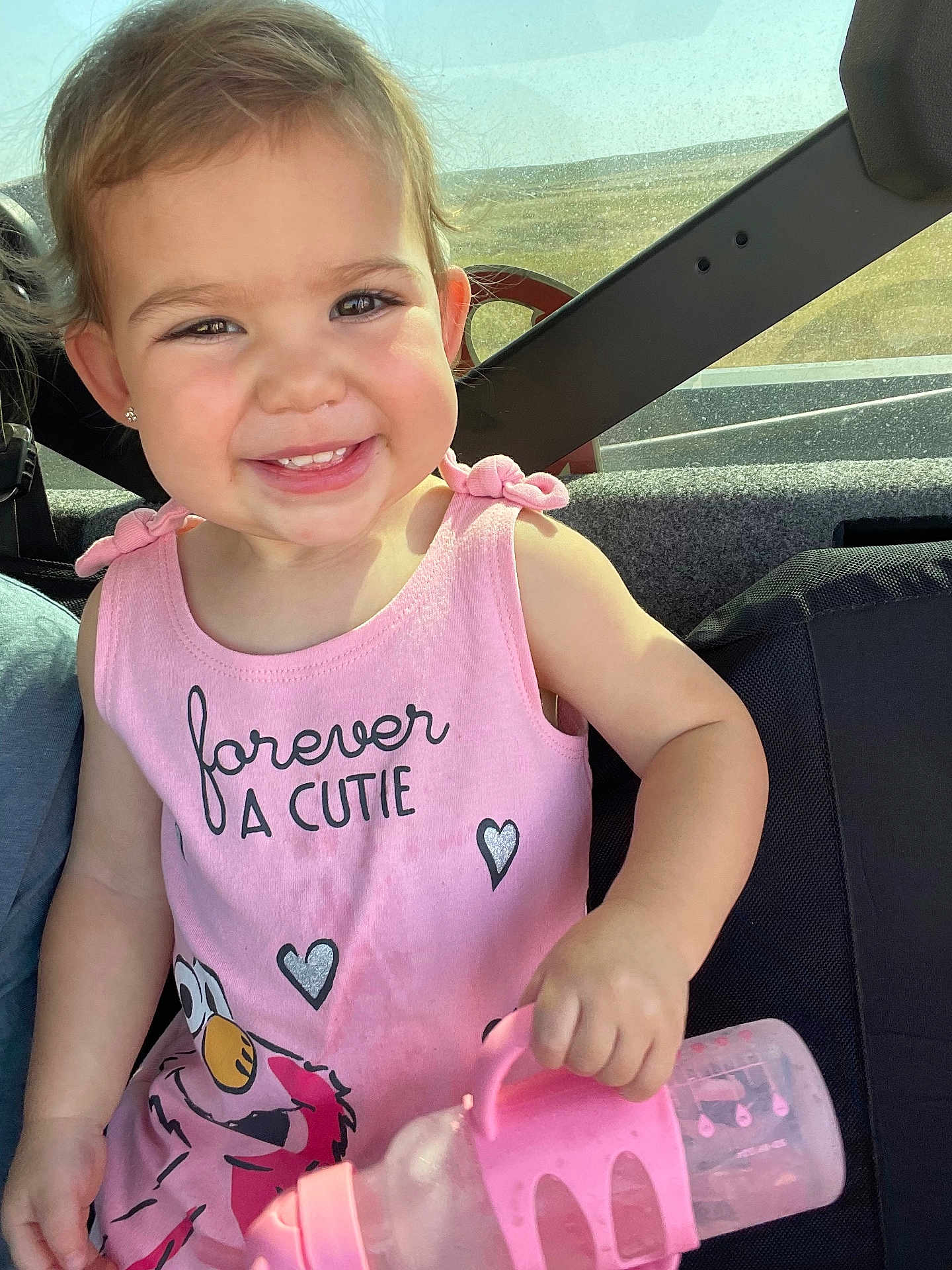 Wrenley is registered to the contest to win money with this photo: toddler, child, smiling, pink_tank_top, water_bottle, car_seat, happy, face, cute, sunlight, earrings, short_hair, vehicle_interior, seatbelt, hand, clothing, portrait, baby, kid, daylight