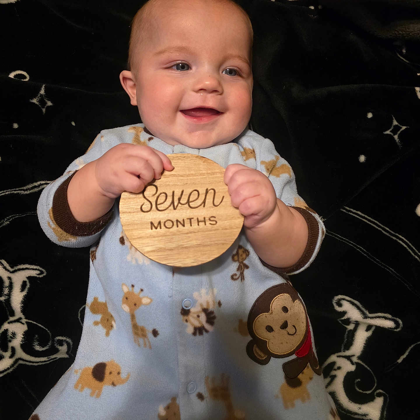 Killian is registered to the contest to win money with this photo: animal_print, baby, blanket, child, cute, elephant, face, giraffe, hands, happy, indoor, infant, lying_down, monkey, onesie, pattern, plaque, portrait, smile, wooden_sign
