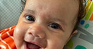 Azael joined the competition — help win amazing prizes! baby, blue_eyes, brown_hair, child, close_up, curly_hair, cushion, face, fleece, happy, indoor, infant, laughing, orange_outfit, portrait, rainbow, seated, smiling, striped_clothing, sun_pattern