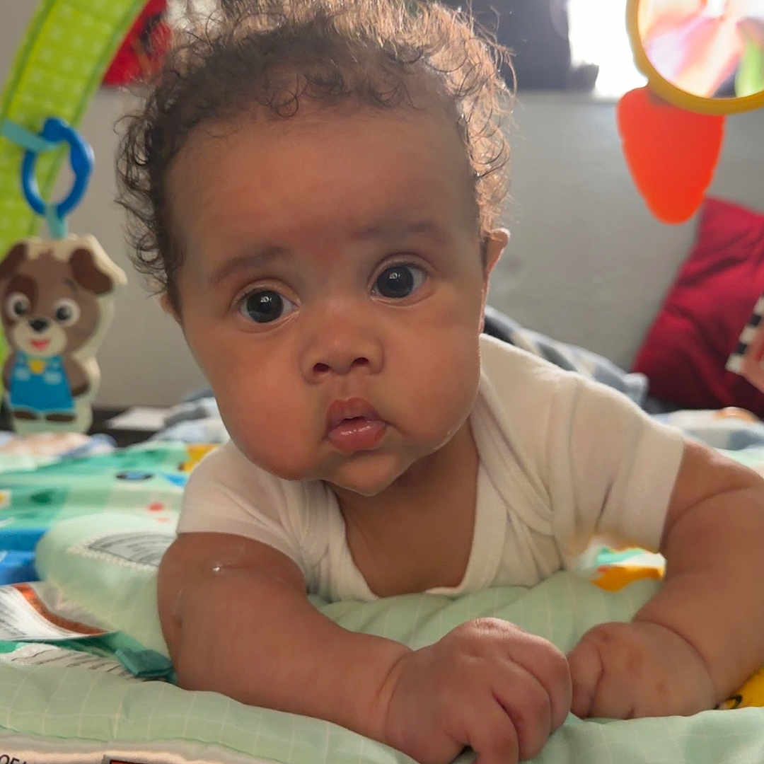 Azael is registered to the contest to win money with this photo: baby, child, infant, tummy_time, play_mat, play_gym, toy, colorful, curly_hair, wide_eyes, portrait, indoors, sitting, lying_down, onesie, hands, arms, nursery, daylight, adorable