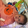 baby, infant, child, portrait, lying_down, wide_eyes, curly_hair, striped_clothing, pumpkin, halloween, blanket, bed, crib, baby_toys, activity_book, colorful, cartoon, nursery, cozy, indoors