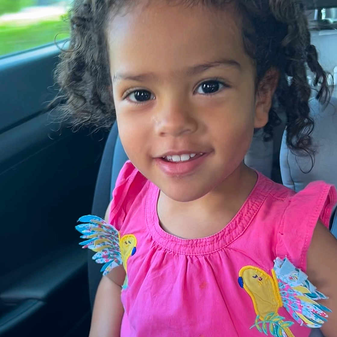 Xiomara a rejoint le concours — aidez-le/la à gagner de superbes lots ! car, car_seat, child, curly_hair, cute, face, girl, hands, happy, interior, parrot_design, person, pink_dress, portrait, seat, seatbelt, smiling, sunlight, window, young