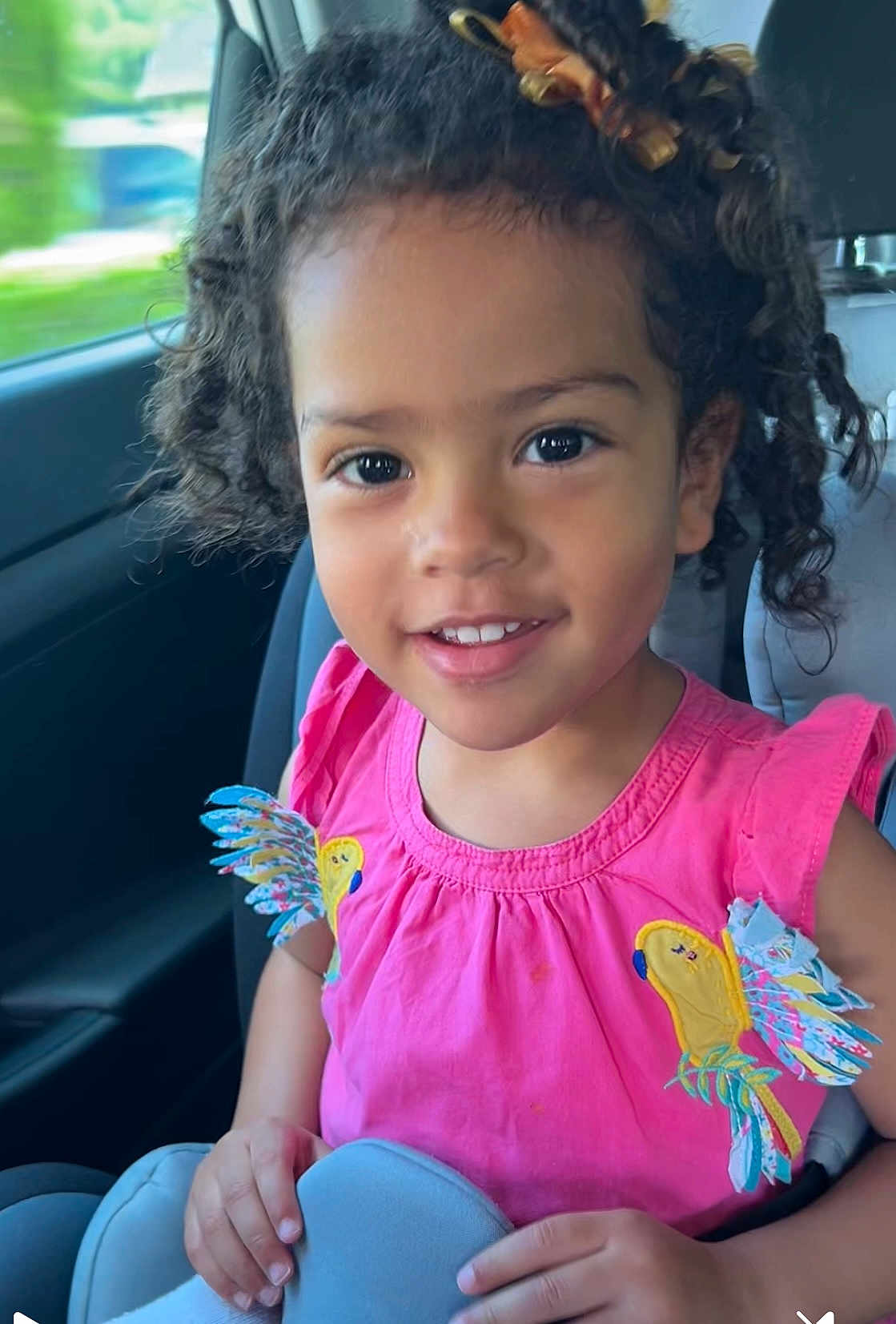Xiomara a rejoint le concours — aidez-le/la à gagner de superbes lots ! child, girl, smiling, car_seat, curly_hair, pink_dress, parrot_design, hands, seatbelt, interior, car, portrait, happy, cute, young, sunlight, window, seat, face, person
