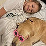 child, dog, sunglasses, bed, blanket, white, smiling, lying_down, indoor, pet, cute, relaxed, pink, accessory, comfort, happy, face, clothing, canine, human